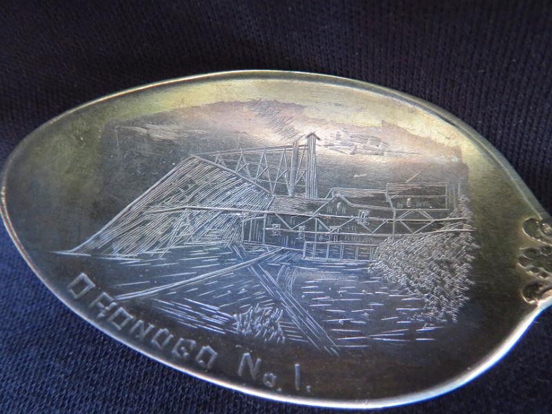 Souvenir Mining Spoon Bowl Marking Oronogo No. 1 Mine, Oronogo, MO.JPG - SOUVENIR MINING SPOON ORONOGO MINE NO 1 ORONOGO MO [Sterling silver spoon, 5 7/8 in. long, engraved mining scene in bowl, bowl marked OROGONO NO. 1, handle front in ornate Versailles pattern introduced in 1888 by Gorham;  It shows an angel holding a musical instrument, maybe pipes or a lyre; reverse is marked with the old lion-anchor-G  hallmark for Gorham, K in a square shape and Sterling; weight 30.9 gms. [Eight miles north of Joplin, Missouri stands the village of Oronogo, originally named Minersville when it sprang up in 1850. According to legend, a miner dug there with his pick and shovel and said, "It's ore here or no go," and so the name Oronogo stuck. Nearby is the site of the richest lead and zinc strike ever made in southwest Missouri. Tom Livingston first discovered the mine before the Civil War, but renegades stole his land while he was away fighting. After the war, they sold it to Granby Mining Company for $50. After almost 80 years of continuous operation, the mine produced $30 million worth of lead and zinc. The Oronogo Circle Mine No. 1, one of the few open-pit mines in the district, stretched 300 feet deep and 600 feet across. The 12-acre unroofed cavern was strip mined at three levels-150 feet, 240 feet, and 360 feet--a flurry of activity with men, trucks, and machinery going up and down the steep inclines. Numerous mills serviced the operation. The largest chunk of pure lead ever found in the district came from the Oronogo Circle. Two flat cars carried it to the World's Fair in Chicago in 1893. After the exhibit ended, the mammoth rock sold for $6,000. A deadly accident occurred at the mine in 1901 when a blast of dynamite detonated prematurely, killing 12 men and wrecking a $50,000 mill. Operations shut down for several months because miners refused to go back to work. Chicago capitalists ran the mine from 1906 to 1914, raking in profits of $3 million. They sold it to the Connecticut Mining Company for $500,000. The ground was so honeycombed with shafts and drifts at four levels that it was no longer safe to work underground. In an ill-fated endeavor to make mining the Circle safer, the new owners closed the old shafts and dismantled 20 small mills. They removed underground support pillars and stripped the entire tract down to 300 feet, using the largest steam shovels available. Then they buried dynamite at the former mill sites and ignited them all at once, causing a blast that almost blew Oronogo off the map. The ground shook so violently that a new mill, built on solid ground, fell into the pit and killed six men. The catastrophe cost the company more than $1 million. Connecticut Mining went out of business, and the mine reverted to its original owner, the Granby Mining Company. For many years, the tract lay idle, although investors made half-hearted attempts to resume strip mining. By the 1970s, the huge water-filled pit had become popular with scuba divers, who liked to explore its depths, yet even that turned out tragically, with the deaths of some of the divers.]